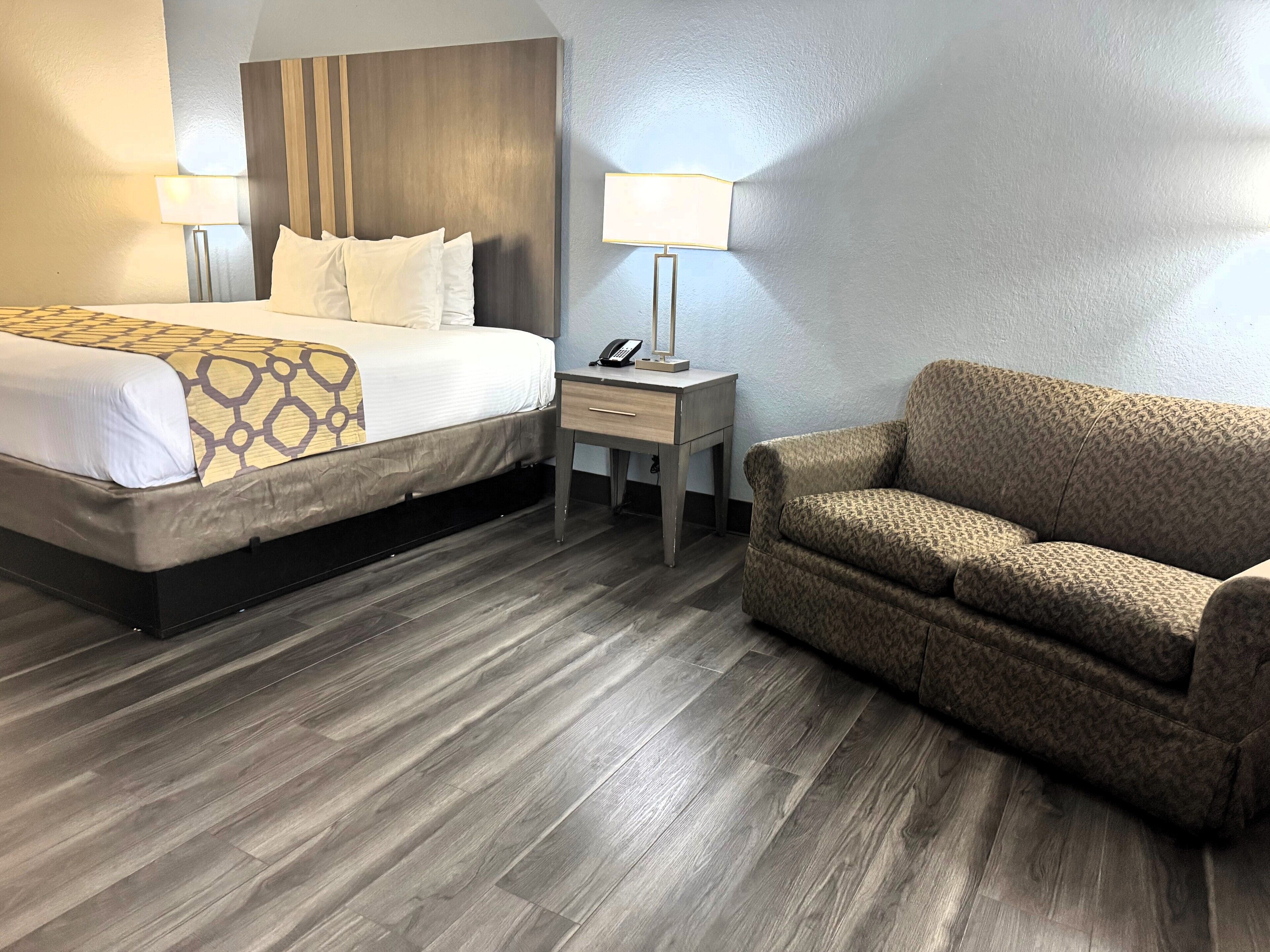 A hotel room with a bed, bedside table, lamp, and a patterned sofa.