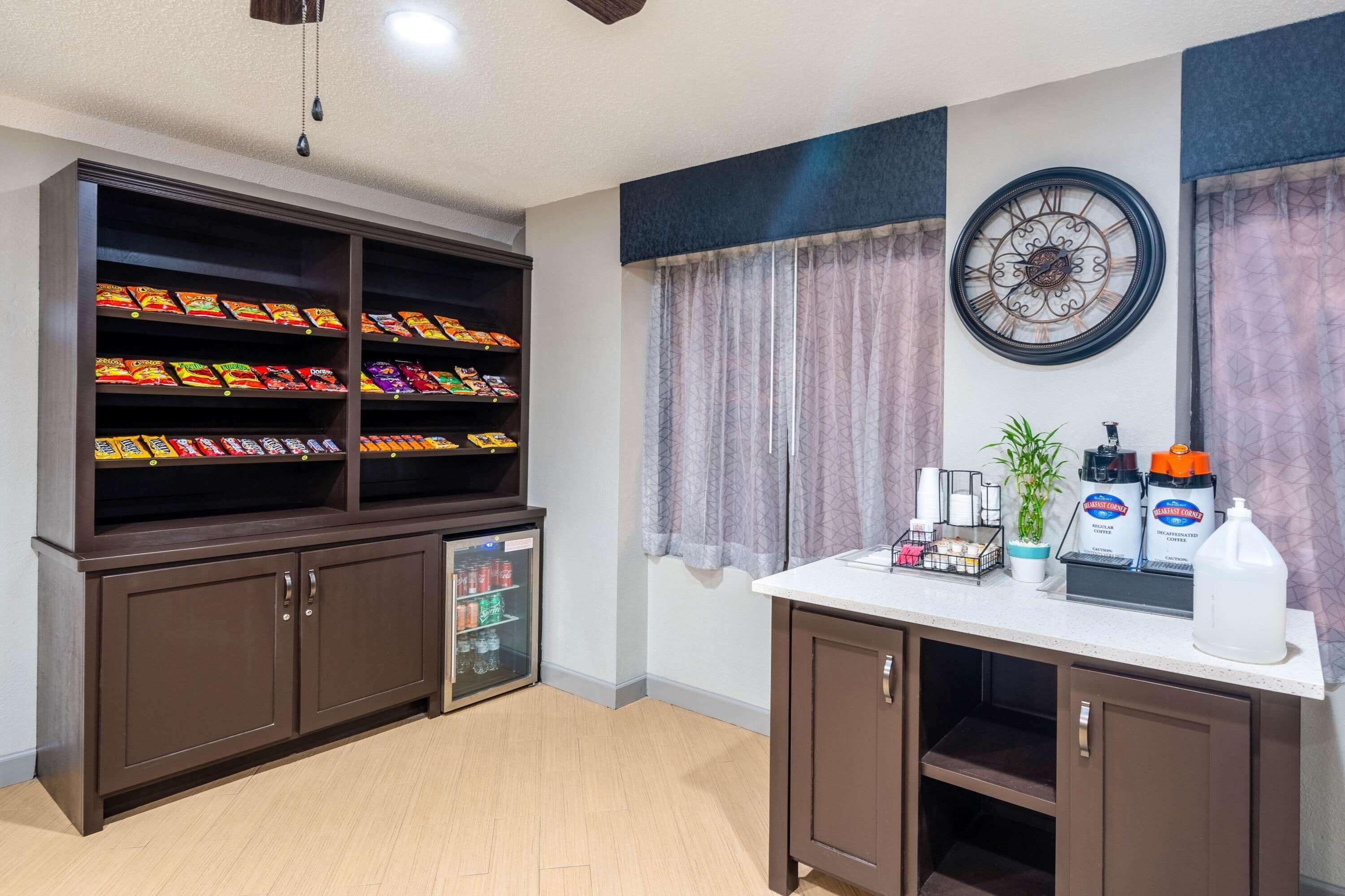 A room with a snack bar featuring a variety of snacks and a refrigerator, a coffee station with a coffee machine, and a clock on the wall.