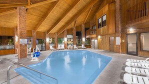 An indoor swimming pool with a wooden ceiling, white lounge chairs, and a brick wall.