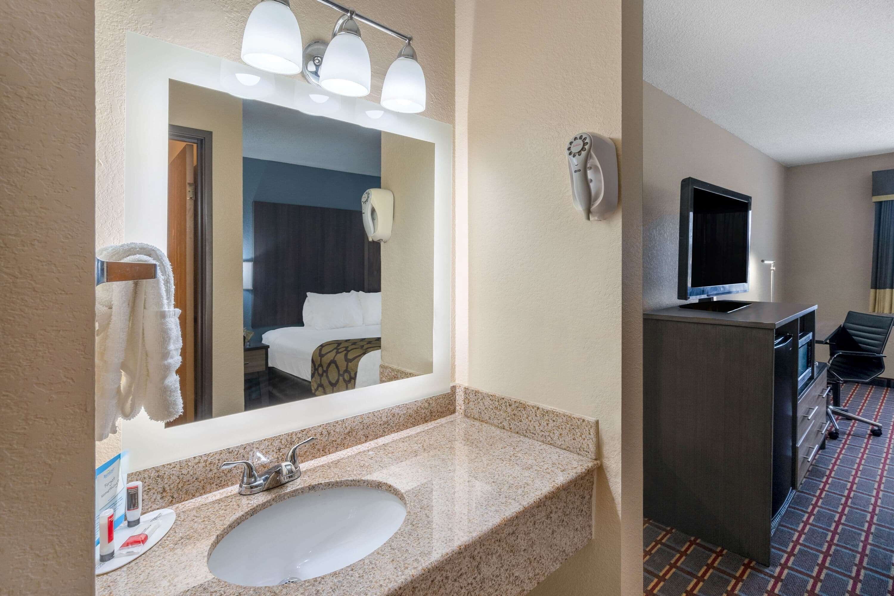 A hotel bathroom with a sink, mirror, towel dispenser, and a television in the adjacent room.