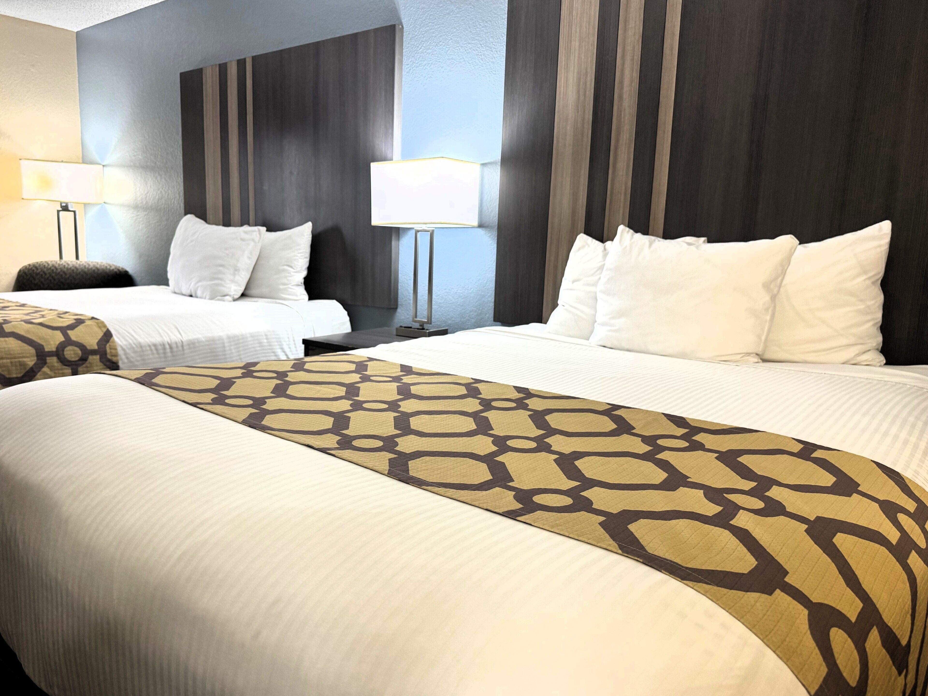 A hotel room with two beds, a large bed with a patterned bedspread, and two smaller beds. There are two bedside tables with lamps.