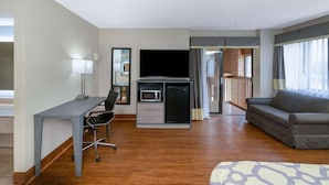 A hotel room with a wooden floor, a desk with a chair, a sofa, a TV, a mini-fridge, and a sliding door leading to a balcony.