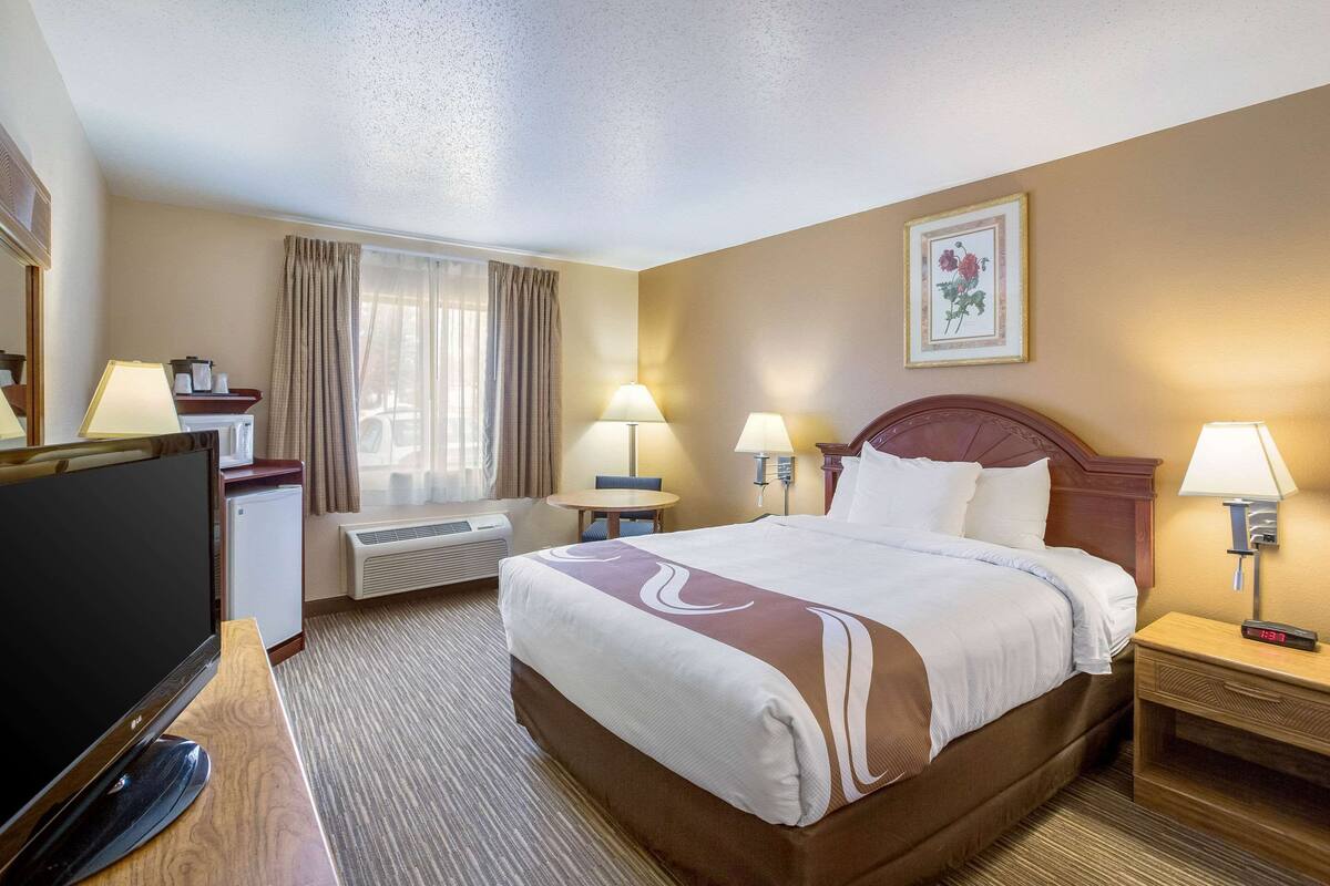 Habitación de hotel con cama, lámparas de noche, un escritorio con televisor, una mesita y una ventana con cortinas.