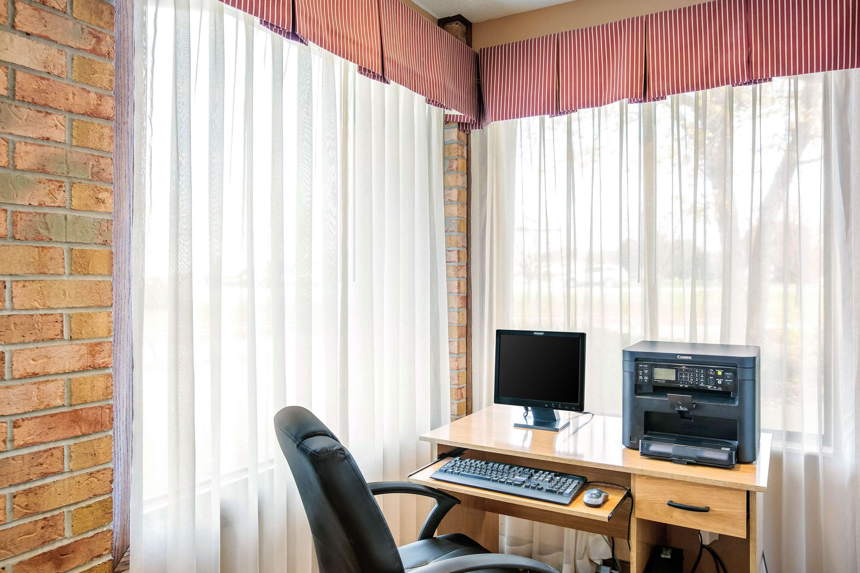 A corner office with a brick wall, a desk with a computer and printer, a chair, and sheer curtains with a striped valance.