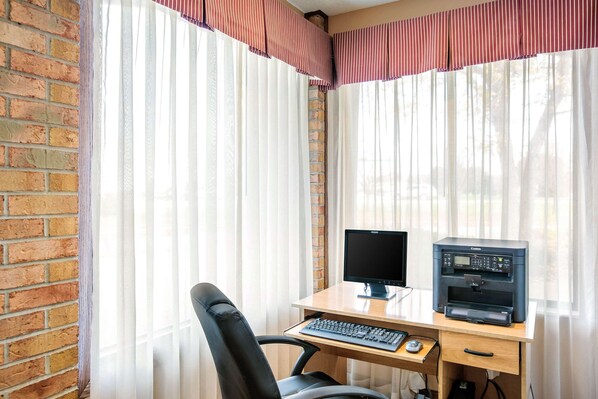 A corner office with a brick wall, a desk with a computer and printer, a chair, and sheer curtains with a striped valance.