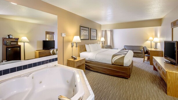 A hotel room with a large bed, a separate sofa, a desk, a TV, a bathroom with a bathtub, and a mirror. - Quality Inn - Storm Lake (Storm Lake)