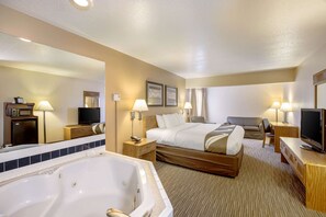 A hotel room with a large bed, a separate sofa, a desk, a TV, a bathroom with a bathtub, and a mirror.