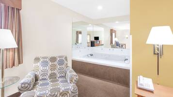 Habitación de hotel con un sillón estampado, un espejo grande, una bañera y una mesita de noche con lámpara y teléfono.