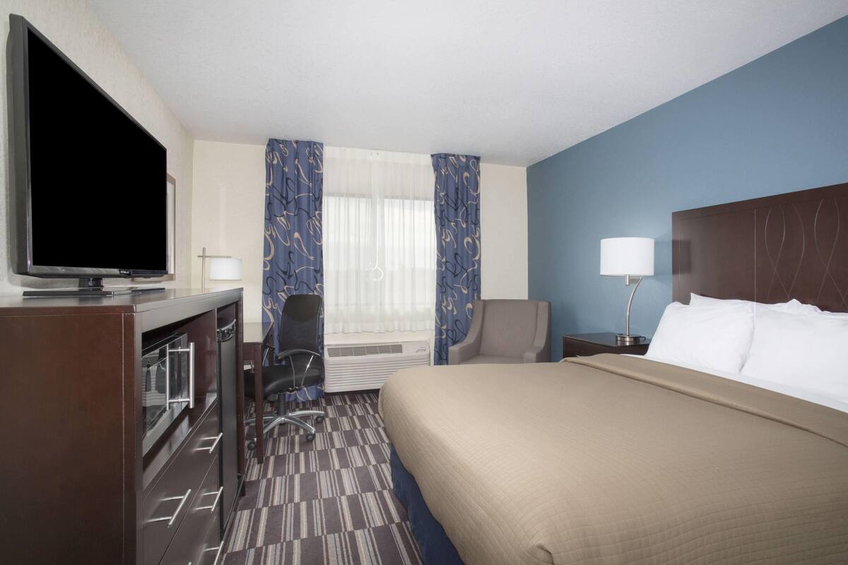 A hotel room with a bed, a desk with a flat-screen TV, a chair, a lamp, and a window with curtains.