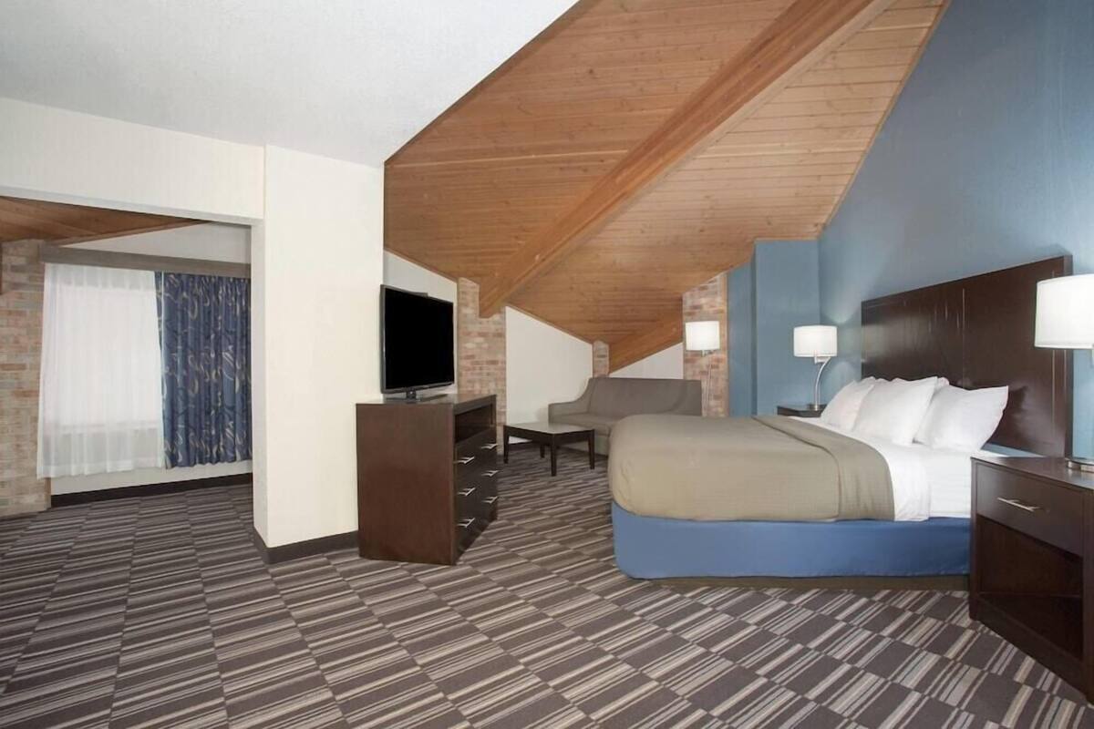 A hotel room with a sloped ceiling, a bed, a desk, a chair, a TV, and a window with curtains.