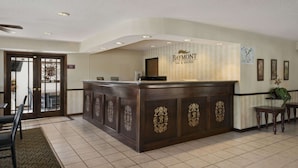 A reception desk with a Baymont Inn & Suites sign, a wooden counter with gold lettering, a patterned rug, and a door with a sun design. - Baymont by Wyndham Whitewater (Whitewater)