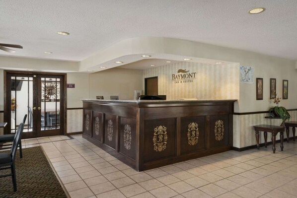 A reception desk with a Baymont Inn & Suites sign, a wooden counter with gold lettering, a patterned rug, and a door with a sun design.
