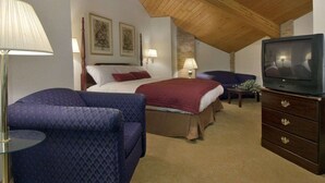 A bedroom with a sloped ceiling, a bed with a red blanket, a blue armchair, a TV, and two wall-mounted lamps. - Baymont by Wyndham Whitewater (Whitewater)