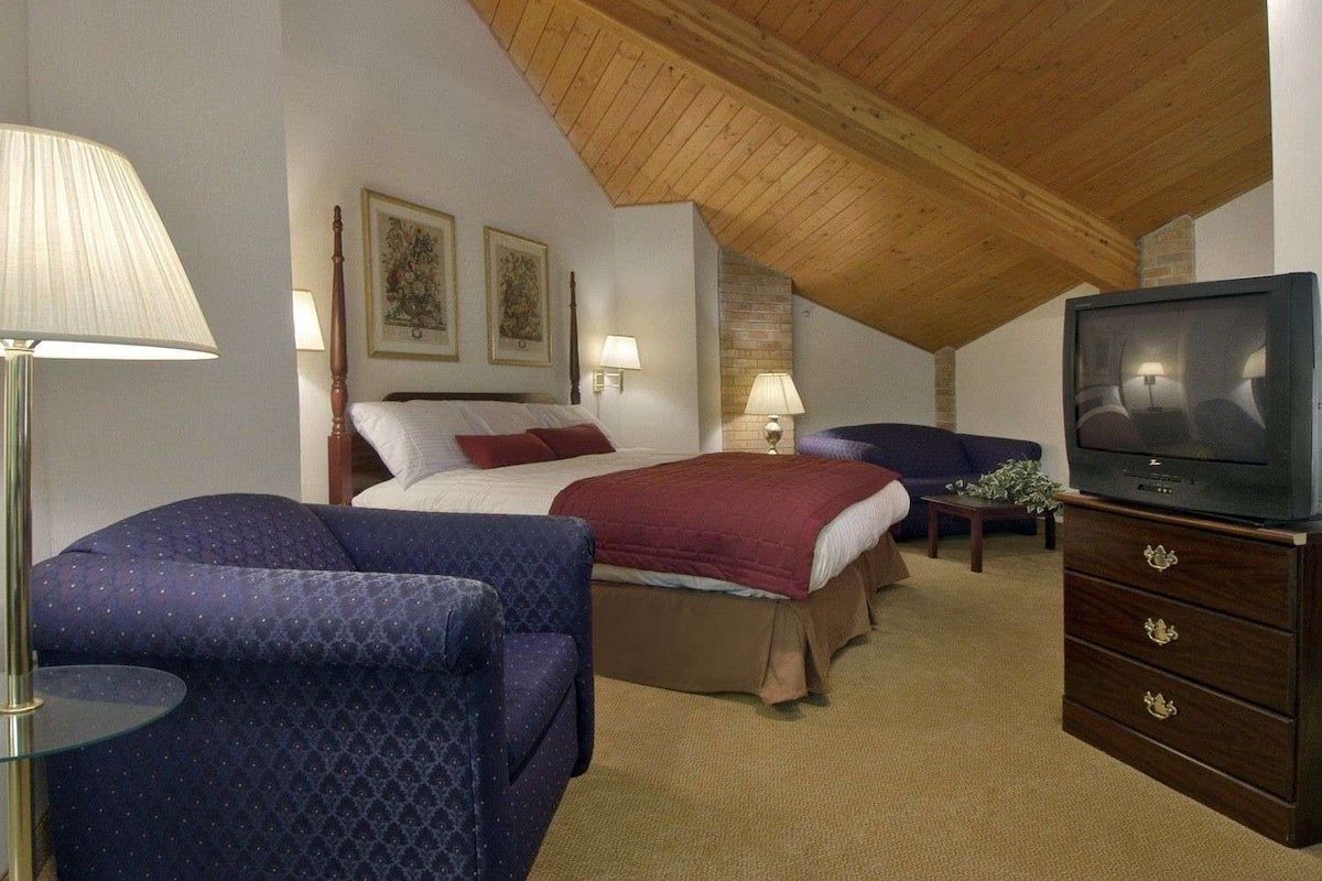 Une chambre avec un plafond en pente, un lit recouvert d’une couverture rouge, un fauteuil bleu, un téléviseur et deux lampes fixées au mur.