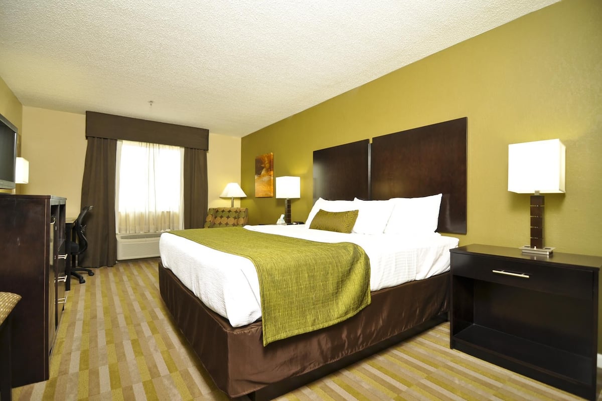 A hotel room with a large bed, two bedside tables with lamps, a desk with a chair, and a window with curtains.