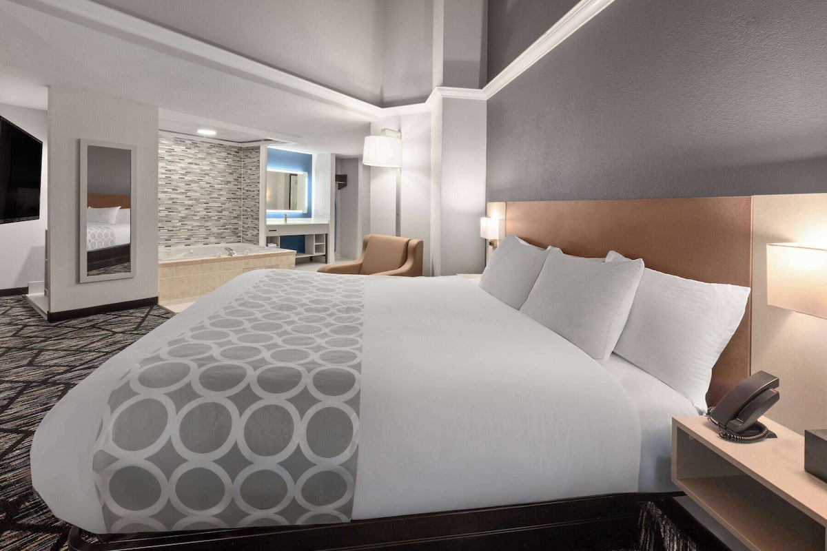 A hotel room with a large bed, a bedside table with a phone, a chair, a bathroom with a mirror and sink, and a patterned carpet.