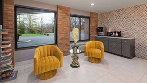 A modern room with two yellow armchairs, a glass coffee table with a decorative elephant sculpture, a brick wall, and a view of a parking lot through large windows.