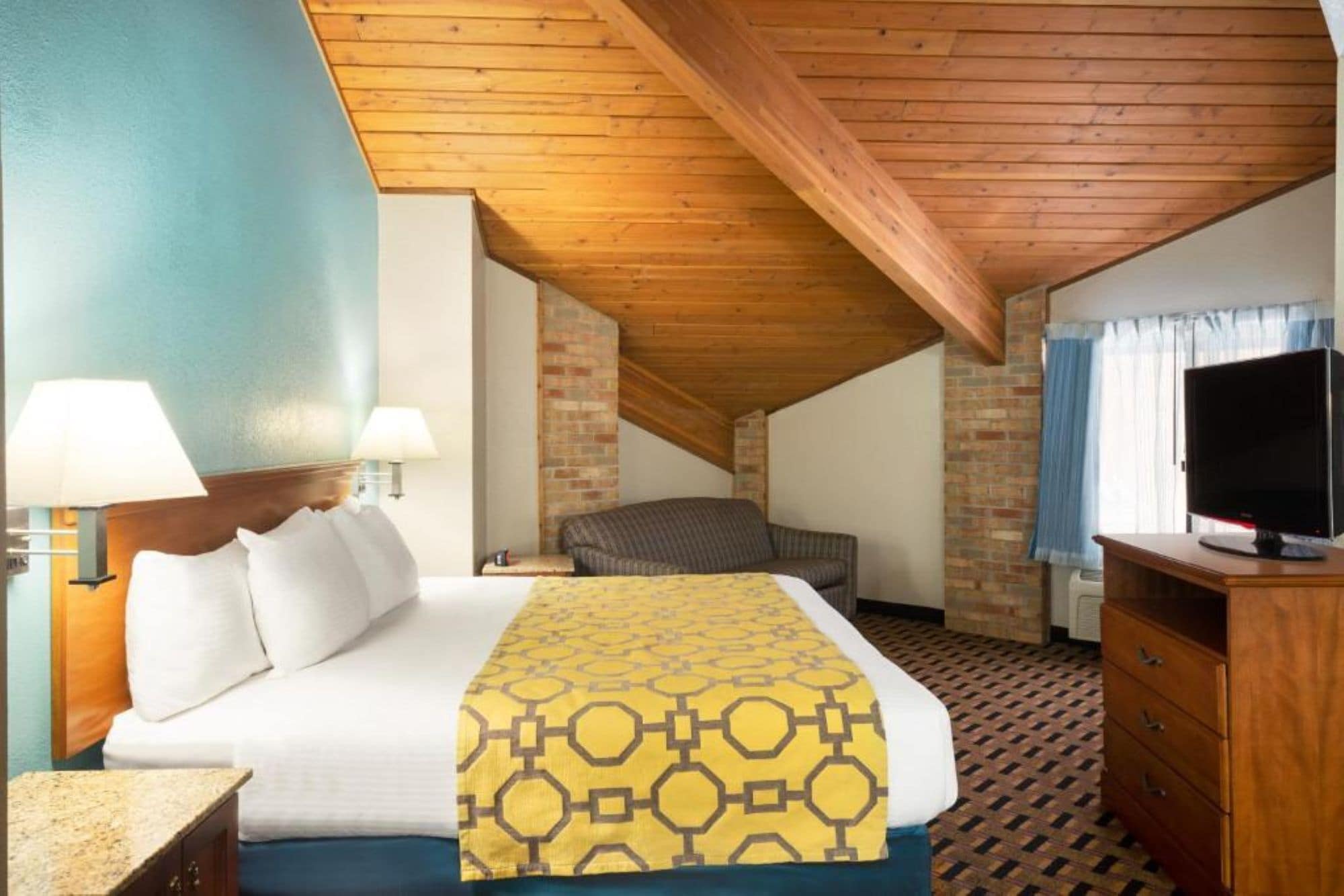 A hotel room with a wooden ceiling, a bed with a patterned bedspread, a television, and a brick wall.