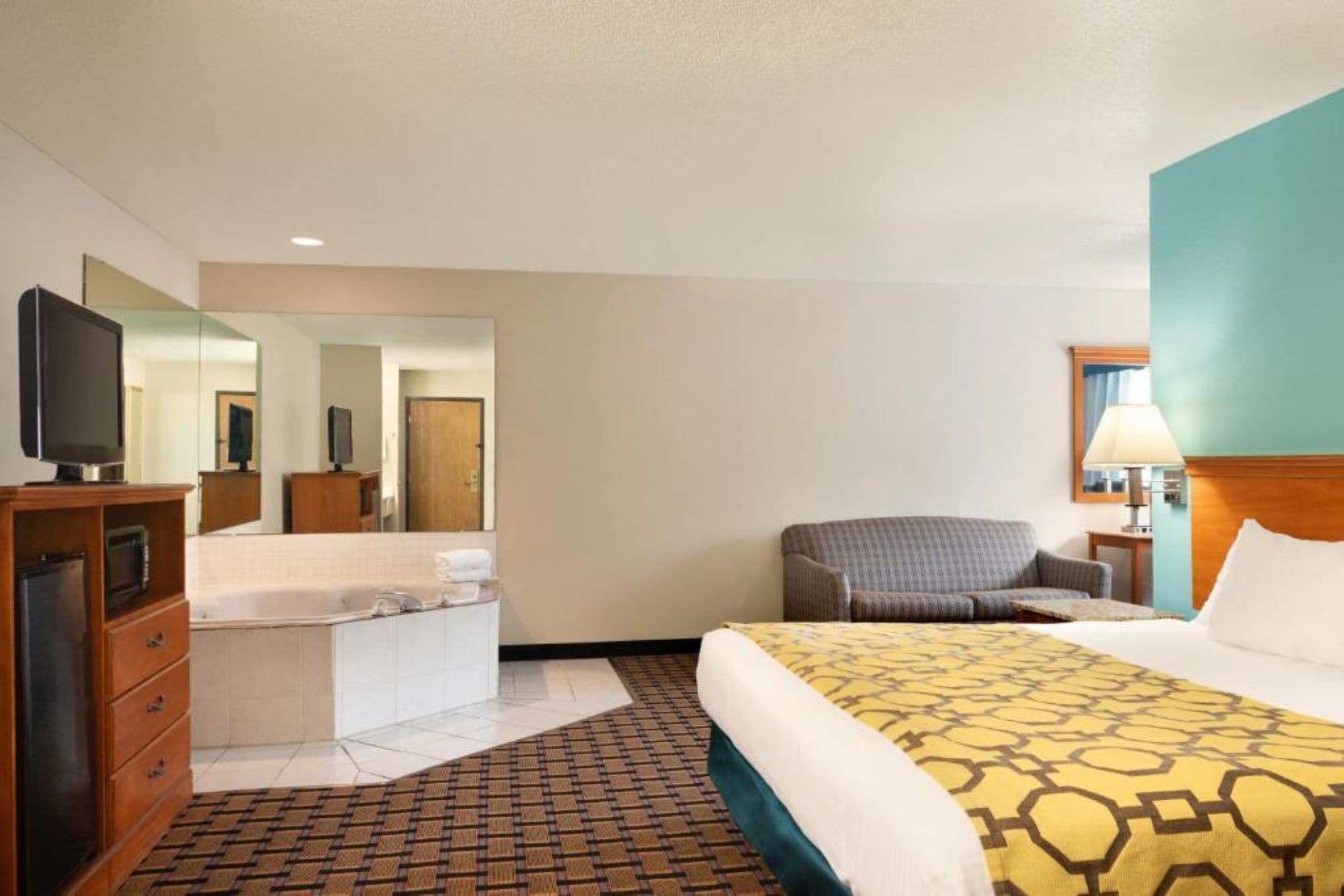 Quarto de hotel com cama, escrivaninha com televisão, cadeira, banheiro com espelho e luminária.