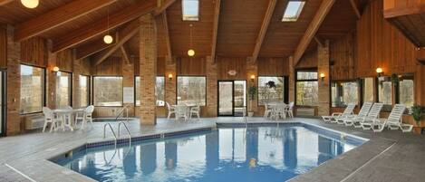 An indoor swimming pool with a wooden ceiling and brick walls, surrounded by lounge chairs and tables.