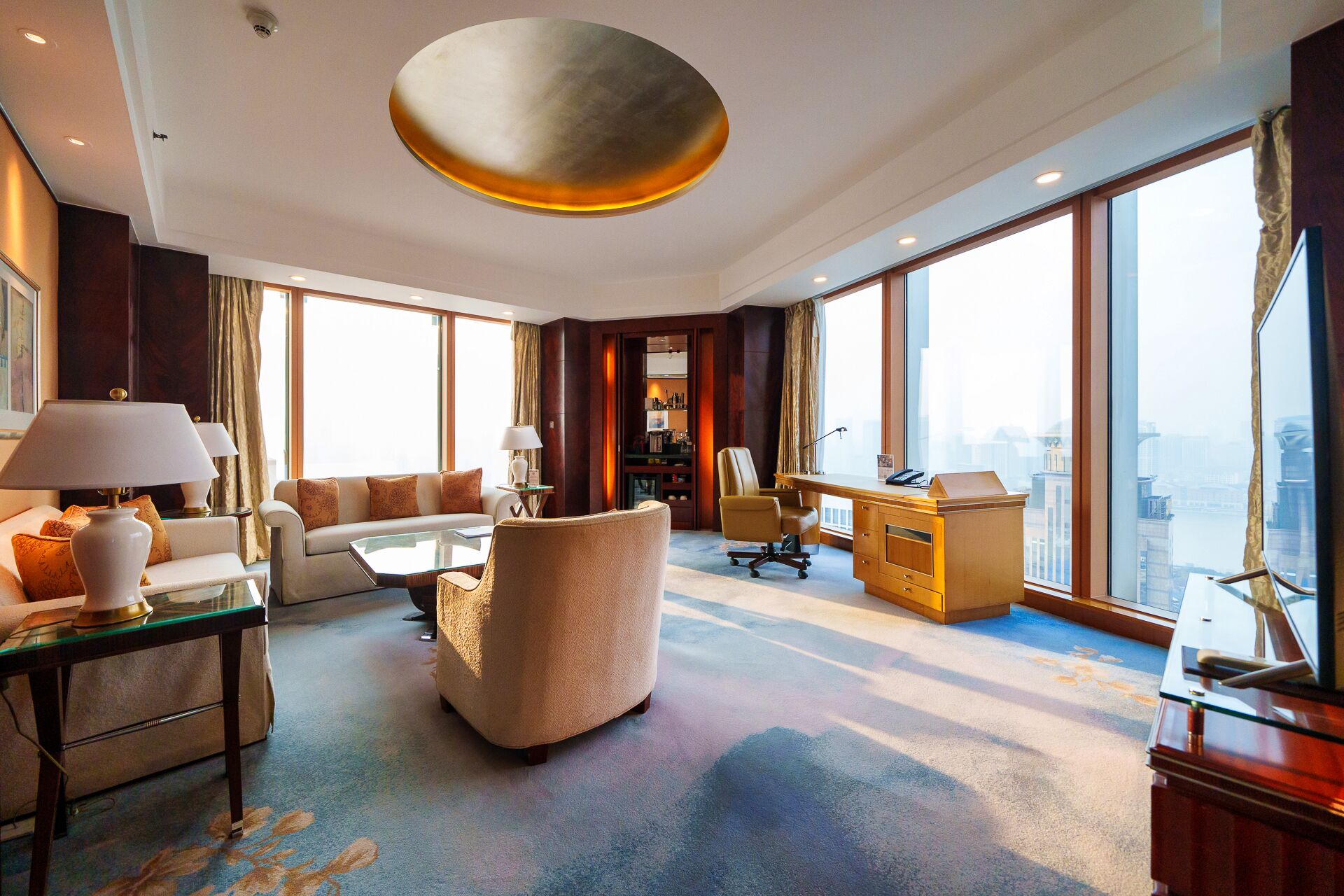 Amplia habitación de hotel con un ventanal grande que ofrece vistas a la ciudad, un sofá cómodo, un escritorio y una lámpara de araña moderna.