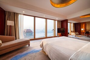 A hotel room with a large bed, a sofa, a TV, and a view of a cityscape through large windows.
