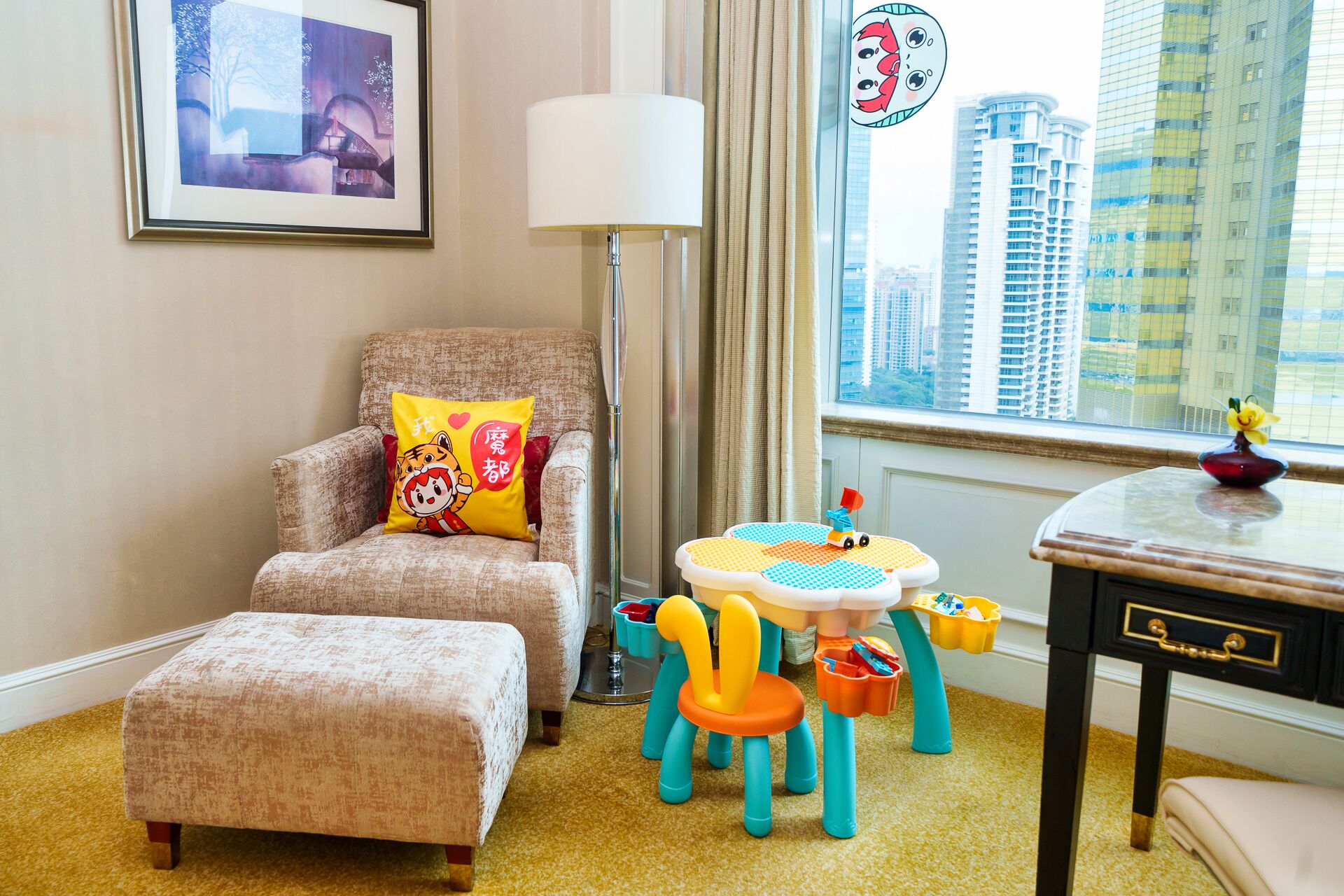 Una habitación de hotel acogedora con un sillón mullido, una mesita con un coche de juguete y vistas a edificios altos a través de la ventana.