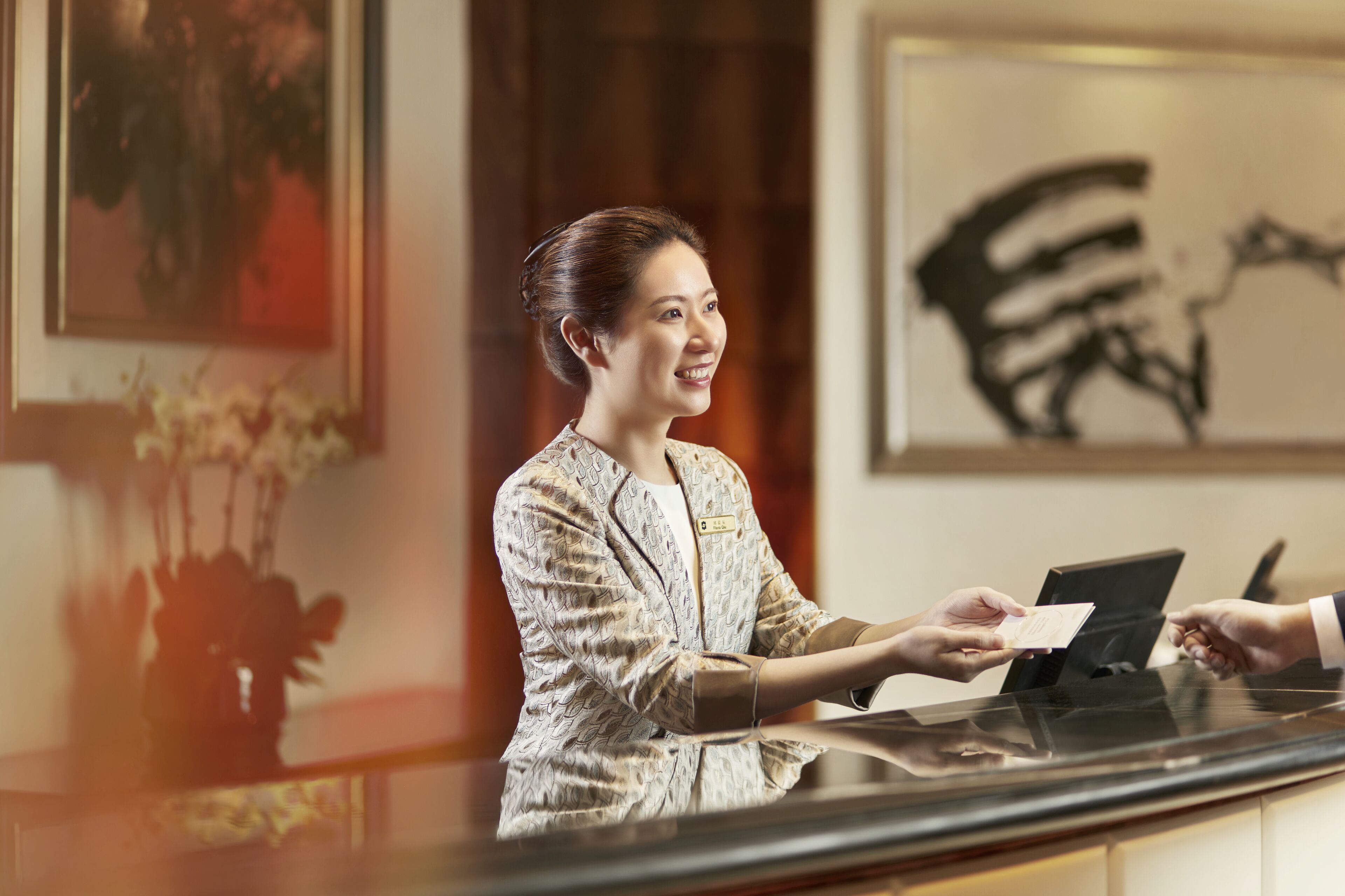 A reception desk with a woman in a patterned blazer, a digital screen, and a framed artwork on the wall.
