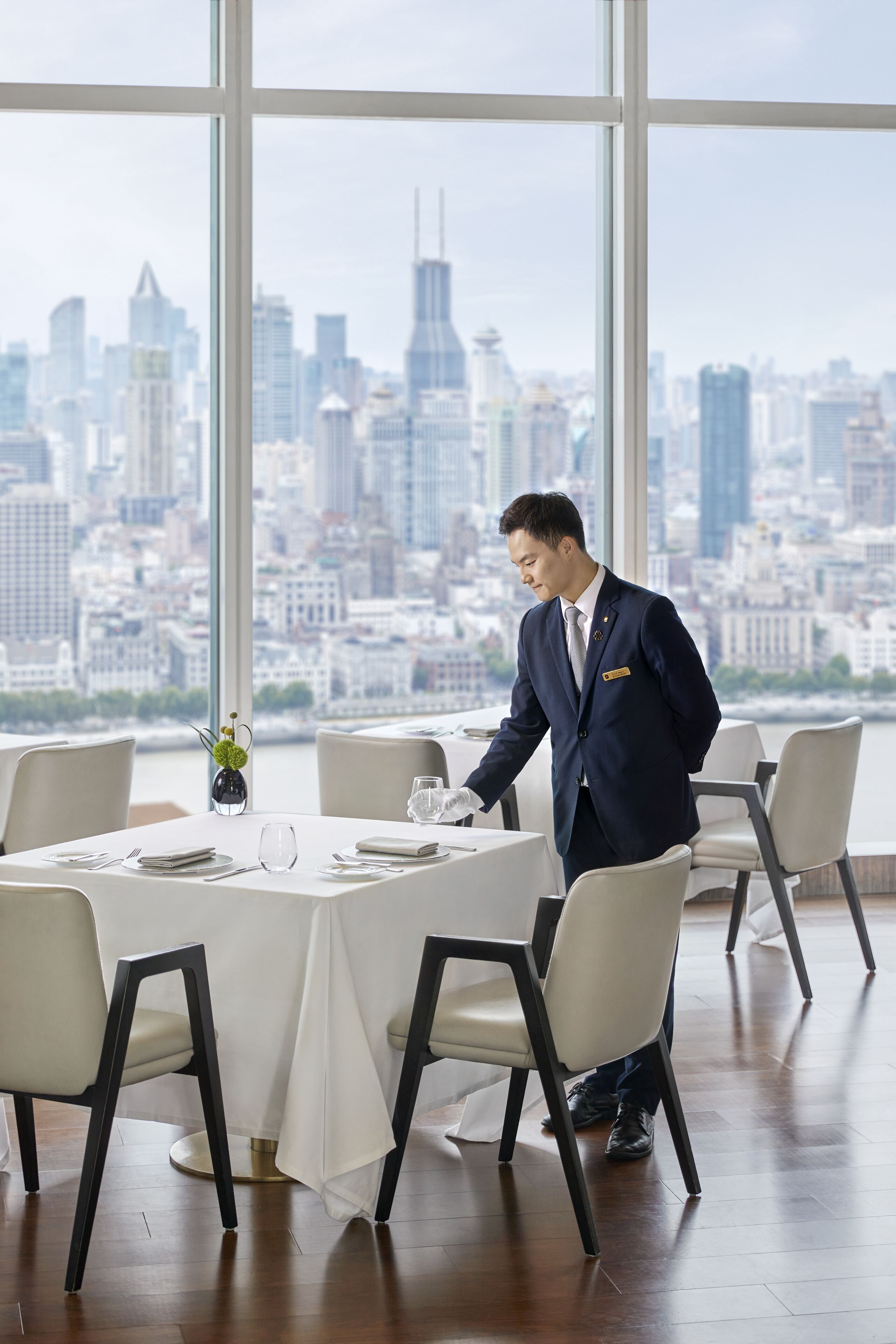 Une salle à manger d’hôtel avec vue sur la ville, un mobilier moderne et un serveur qui s’occupe d’une table.