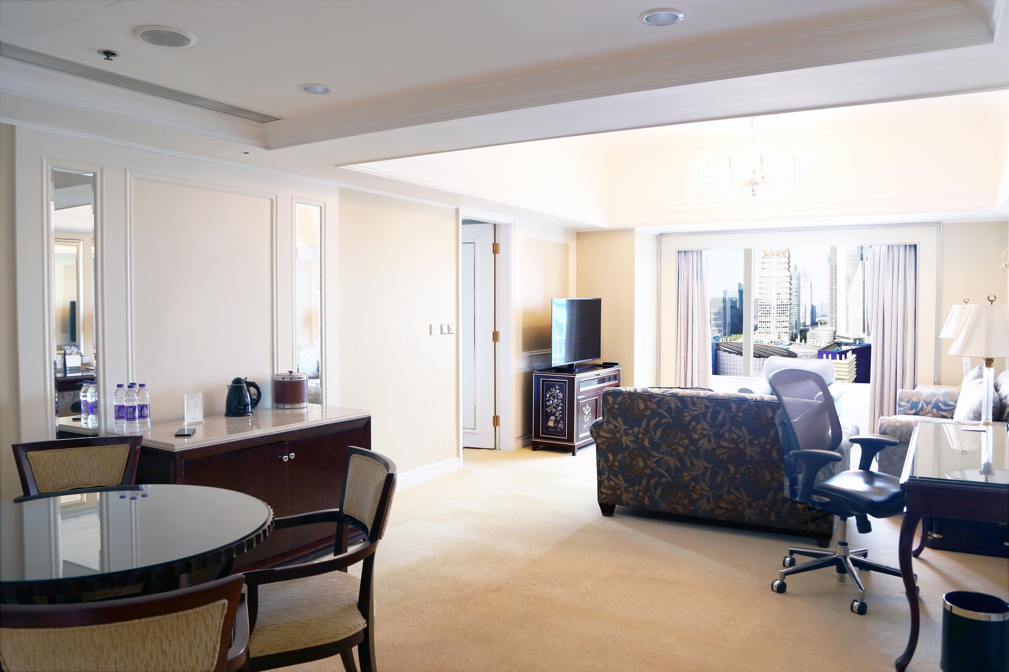 A hotel room with a dining table, chairs, a sofa, a TV, and a city view.