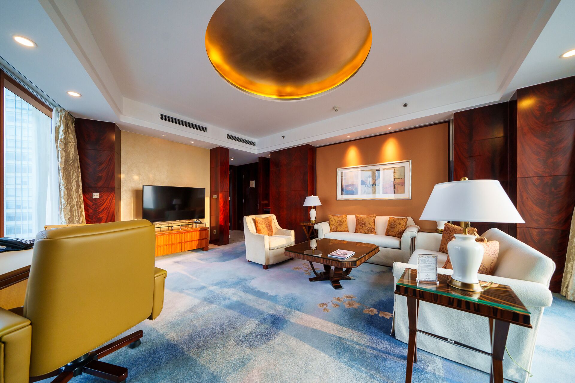 A modern hotel room with a large flat-screen TV, comfortable seating, a coffee table, and a large window providing a view of the city.