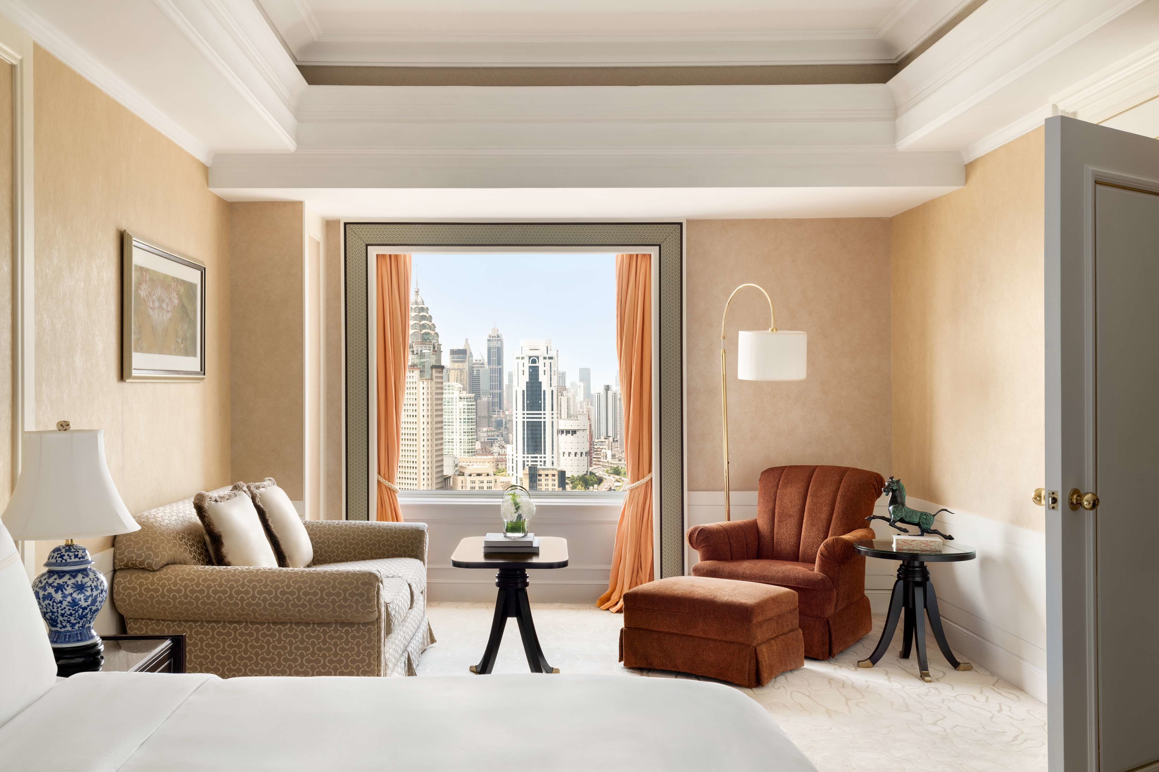 A hotel room with a large window offering a city view, a bed, a sofa, a chair, and a small table with a lamp.