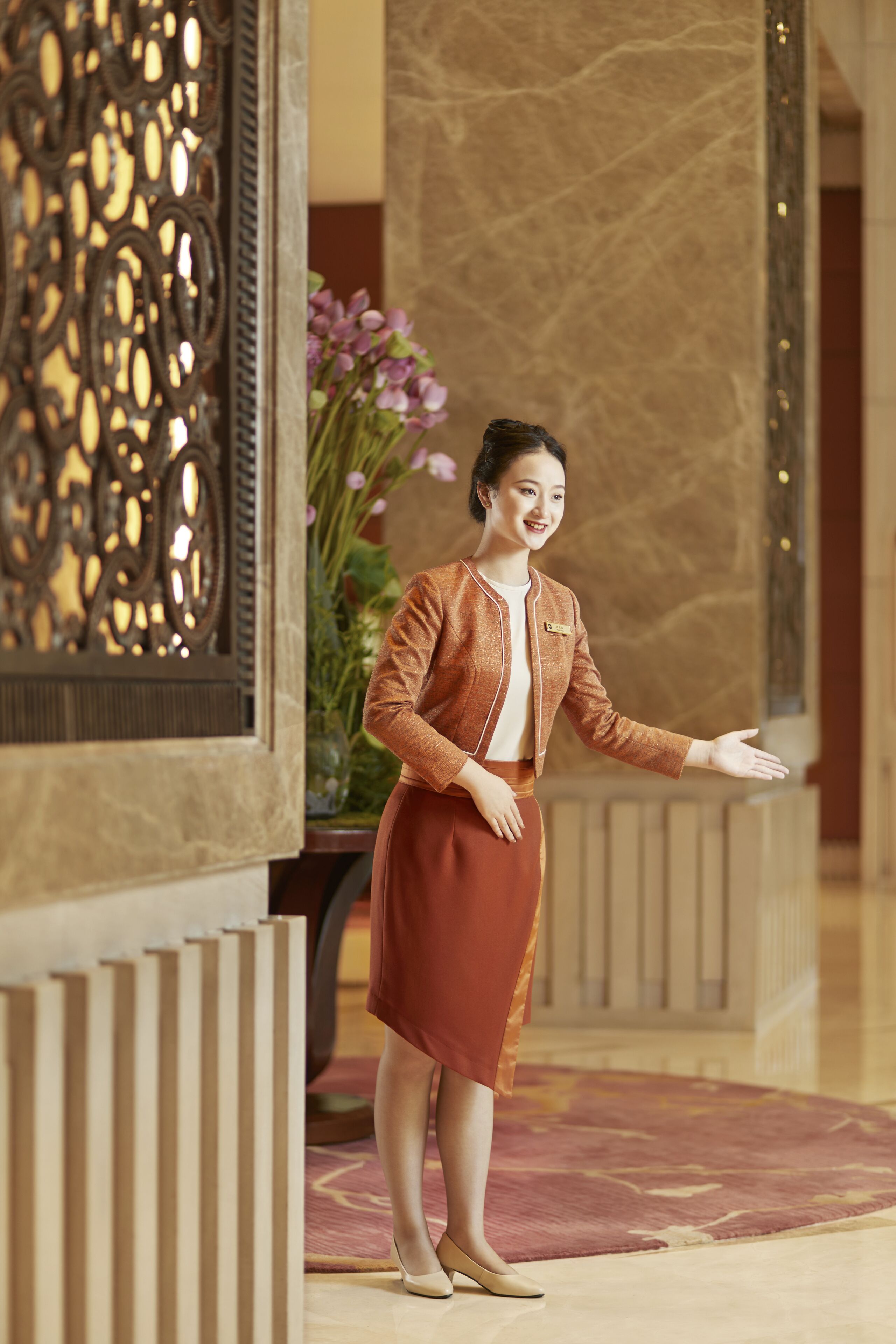 A person standing in a lobby with a decorative panel, a vase of flowers, and a patterned carpet.