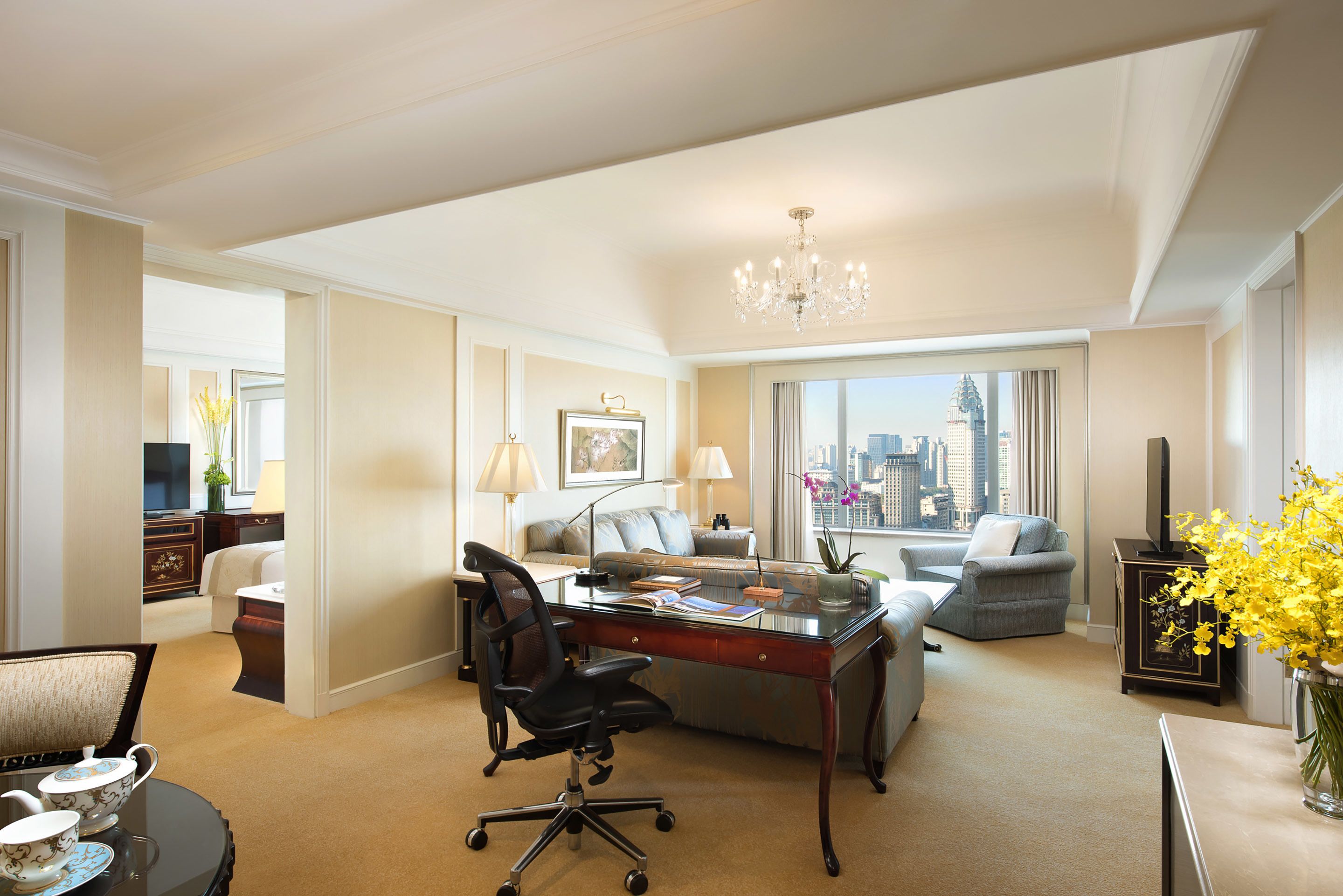 A hotel room with a desk, office chair, sofa, and a large window offering a city view.