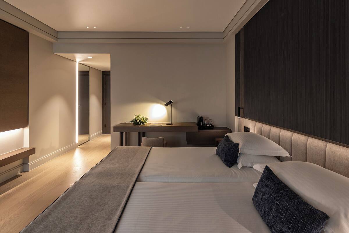 A modern hotel room with a large bed, a desk with a lamp, and a dark wood panel on the wall.