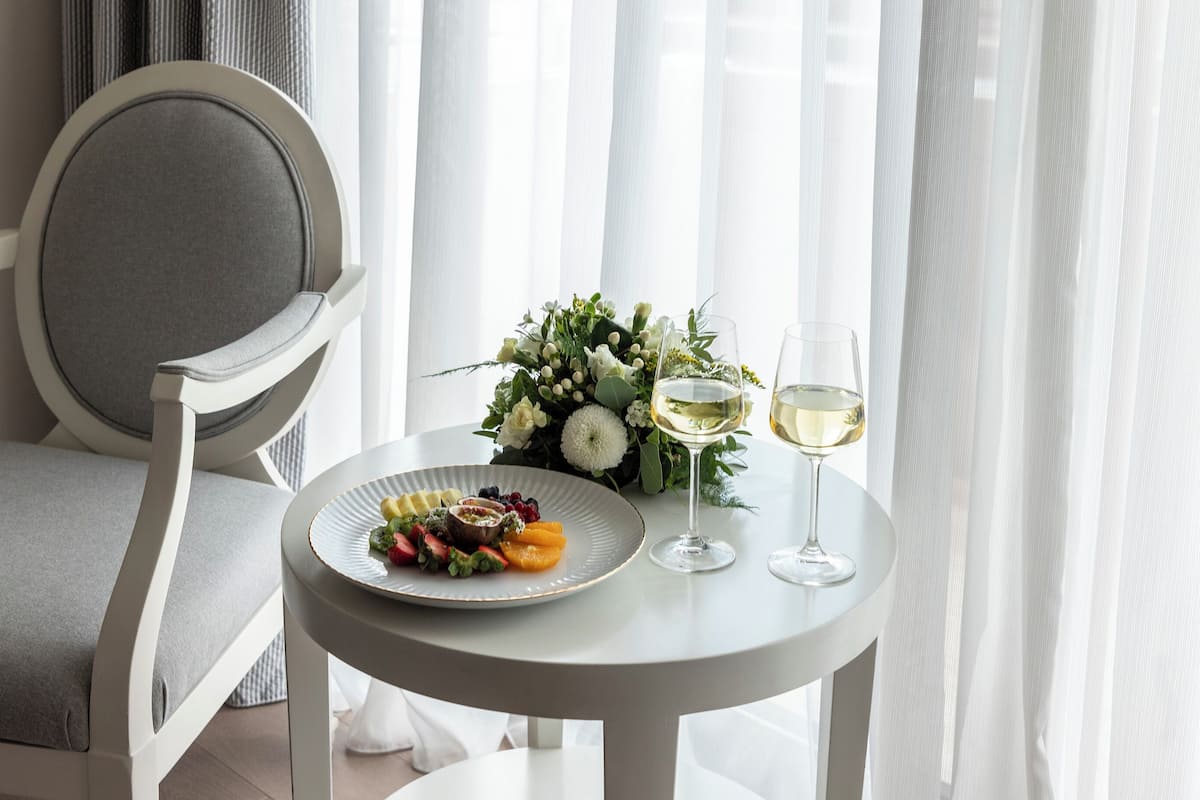 A dining table with a plate of fruit and a glass of white wine, a floral arrangement, and a white chair.