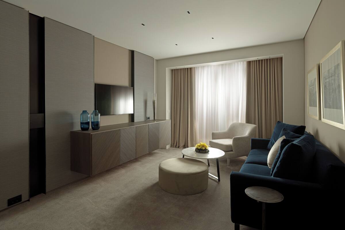 A modern hotel room with a sofa, a coffee table, a TV, and a large window with curtains.