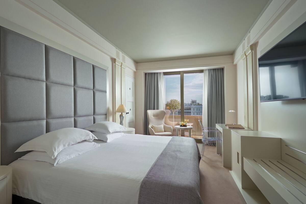 A hotel room with a large bed, a chair, a desk, and a view of the city through the window.