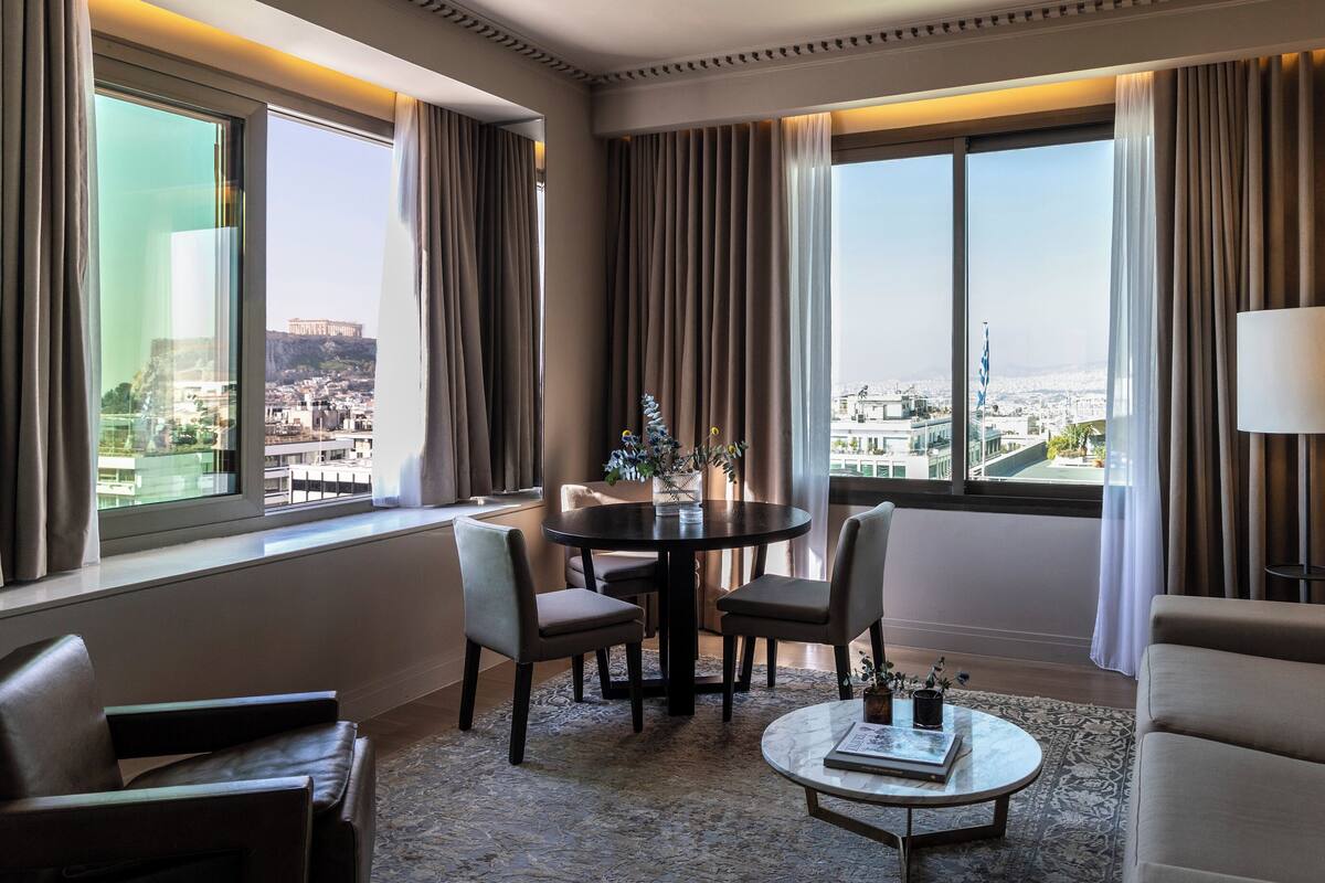 A hotel room with a city view, a round table with chairs, a sofa, and a lamp.