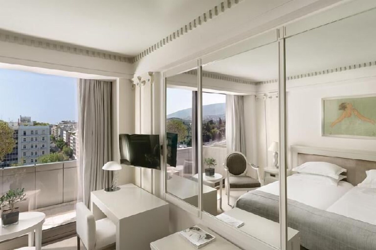 A hotel room with a large bed, a desk with a chair, a television, and a balcony with a view of the city.