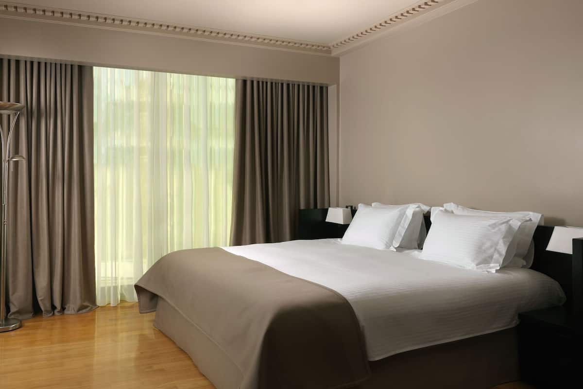 A hotel room with a large bed, a nightstand, a lamp, and a view of the outdoors through sheer curtains.