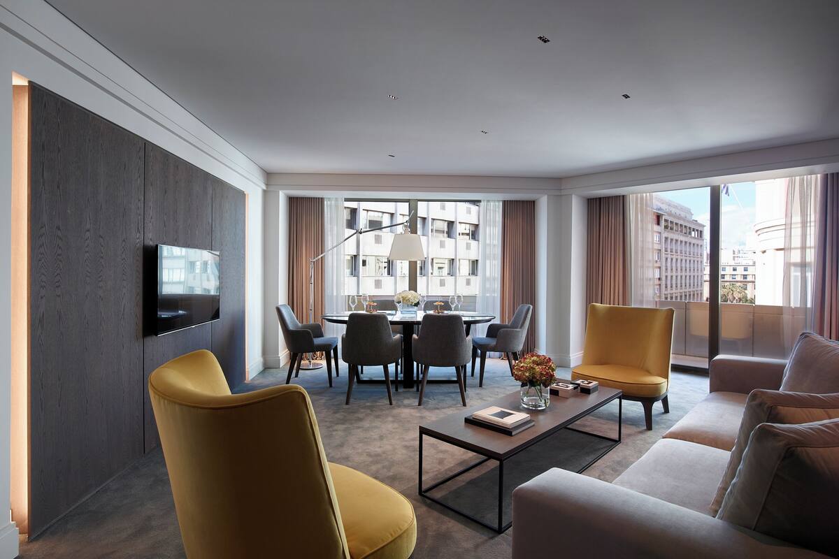 A modern hotel room with a flat-screen TV mounted on the wall, a dining area with a table and chairs, a sofa, and a coffee table with a vase of flowers.