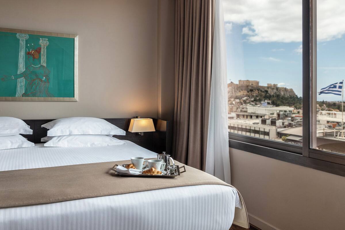 A hotel room with a bed, a painting, a nightstand, a lamp, and a tray with croissants and cups.