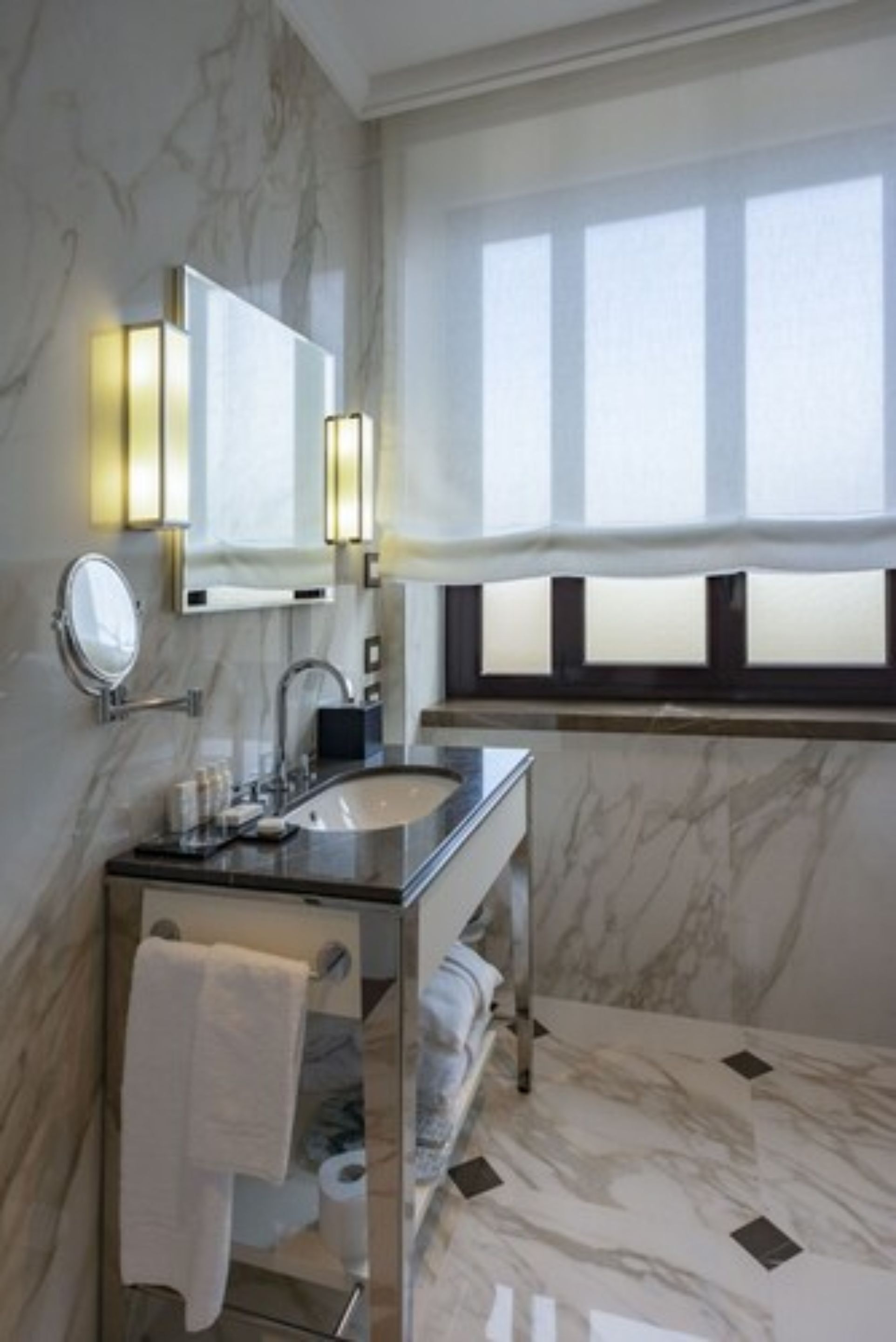 A modern bathroom with a marble sink, a glass vanity, a mirror with wall sconces, and a window with sheer curtains.