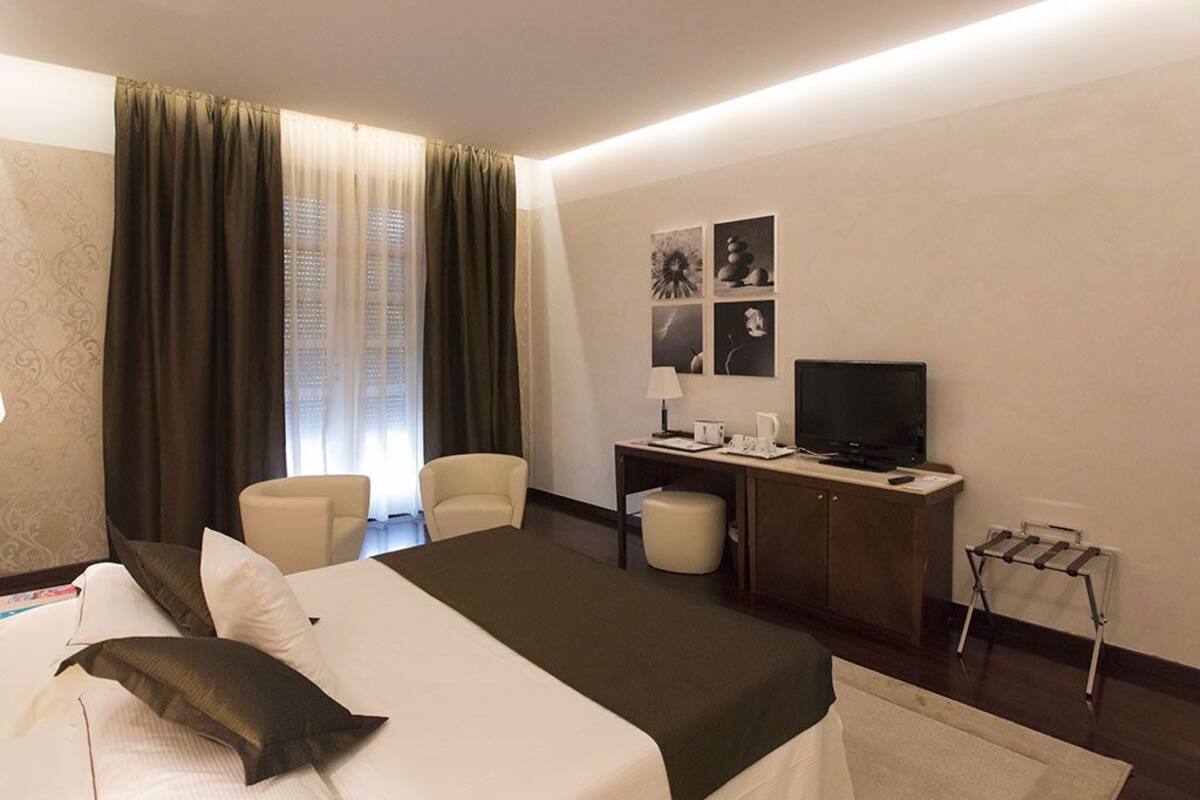 Ein Hotelzimmer mit einem Bett, einem Schreibtisch mit Fernseher, einem Sessel und einem kleinen Tisch mit Lampe.
