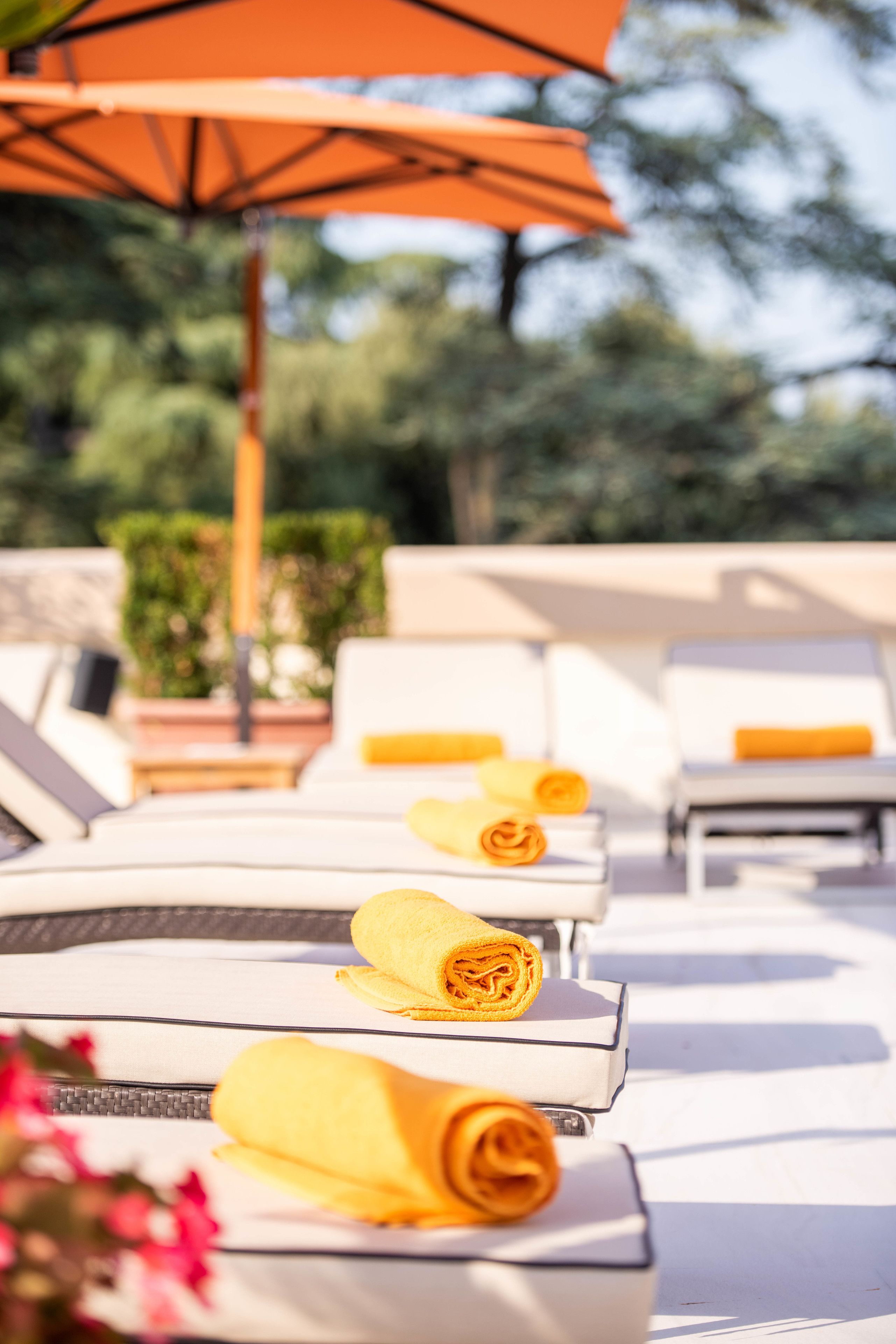 A row of sun loungers with yellow towels, an orange umbrella, and a view of greenery in the background.