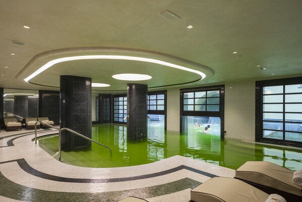 An indoor swimming pool with a green water feature, surrounded by a tiled floor and large windows.