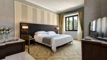 Habitación de hotel con cama, mesitas de noche, televisión, ventana con cortinas y vistas al exterior con vegetación.