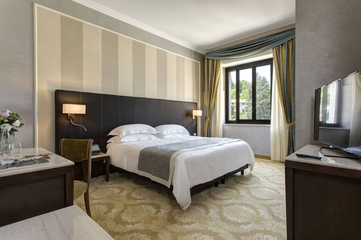 Ein Hotelzimmer mit Bett, Schreibtisch, Stuhl, Fernseher und einem Fenster mit Vorhängen.