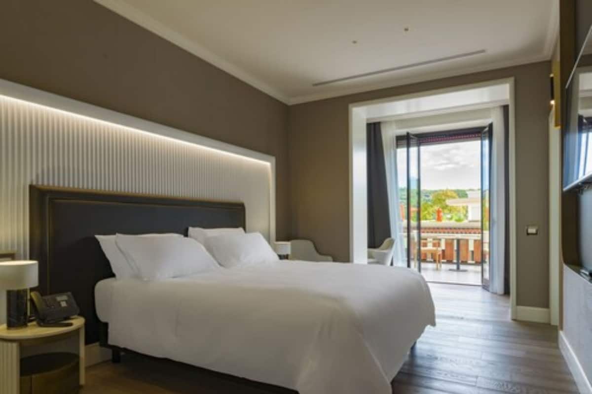 Ein modernes Hotelzimmer mit einem großen Bett, einem Nachttisch mit Lampe und Blick ins Freie durch eine Glastür.